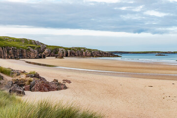 Ceannabeinne Beach shines under a clear summer sky. Crystal-clear waters, golden sand with a heart drawn on it, and lush green grass create a pristine and romantic coastal escape.

