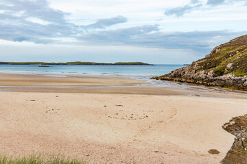 Ceannabeinne Beach shines under a clear summer sky. Crystal-clear waters, golden sand with a heart drawn on it, and lush green grass create a pristine and romantic coastal escape.

