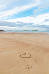 Ceannabeinne Beach shines under a clear summer sky. Crystal-clear waters, golden sand with a heart drawn on it, and lush green grass create a pristine and romantic coastal escape.

