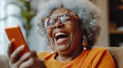 Happy mature old 60s woman holding smartphone using mobile phone app for video call, laughing while watching funny video, feeling excited winning online lottery bid on cellphone sits on couch at home