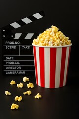 Movie night essentials with a striped popcorn bucket and film clapperboard on a black background. Cinema snack concept for entertainment and film industry.