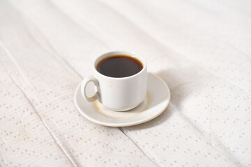 White ceramic cup filled with dark coffee rests on light patterned fabric. Bright lighting highlights coffees rich color, creating cozy atmosphere. Flat perspective emphasizes contrast