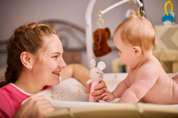 Smiling adult mother with brown hair plays with joyful baby girl on changing table. Soft lighting highlights calm, warm nursery ambiance, and colorful toy
