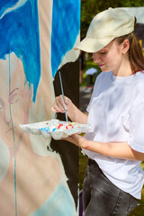 Young female artist with fair hair wearing cap paints large canvas outside. Blue dominates artwork....