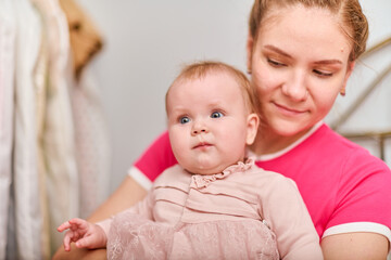 Young mother with light brown hair, holding baby girl. Woman wears pink top, baby in light dress. Warm emotions, indoor environment, bright lighting