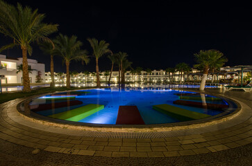 Tranquil resort scene featuring illuminated pool with colorful loungers. Palm trees line poolside against clear night sky. Warm lighting highlights resort architecture