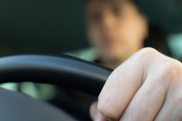 The driver's left hand holds the steering wheel against the background of a blurred face in the car interior