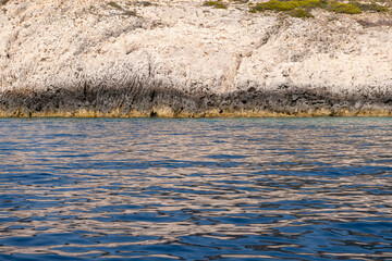 A rugged limestone cliff meets the calm blue sea along the coastline of a Greek island under soft sunlight.