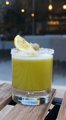 A stylish glass of green juice with a salted rim, lemon slice, and olives, set on a wooden table in an open café space. A casual and refreshing concept perfect for modern food and drink photography.