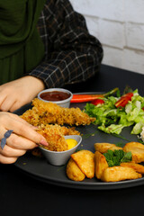 Side view of a woman enjoying a plated meal with crispy fried chicken strips, golden potato wedges, fresh vegetable salad, and dipping sauces on a black plate, against a modern dining background