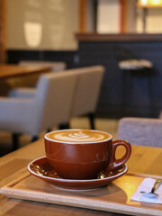 Close-up of a freshly prepared latte served in a deep brown ceramic cup and matching saucer on a natural wooden table in a contemporary coffee shop interior. Soft bokeh captures the blurred