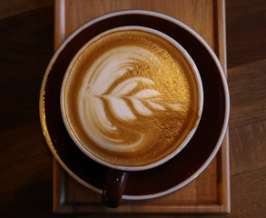Top-down view of a creamy latte with artistic foam leaf design, served in a brown ceramic cup and saucer on a wooden tray. Ideal for elegant flat-lay food photography, coffee blogs, or minimal café