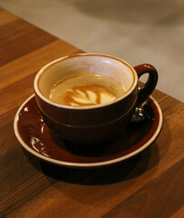 Close-up view of a rich latte served in a brown ceramic cup and matching saucer on a natural wooden table. Creamy white foam art swirls gracefully atop a smooth espresso base, illuminated by warm