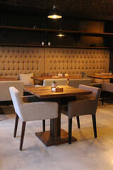 Empty modern café interior featuring a solid wooden dining table flanked by two upholstered chairs. The tufted banquette bench against a textured wall forms a cozy seating nook. Ambient 