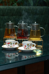 Set of three colorful tea pots with steaming tea cups served on a glass table, cozy warm beverage scene with herbal and black teas, steaming aroma rising, ideal for winter evenings, casual gathering