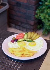 Delicious breakfast plate featuring sunny-side-up eggs, crispy fries, pickles, olives, and tomatoes, beautifully served on a white plate on a clear glass table, bathed in natural sunlight