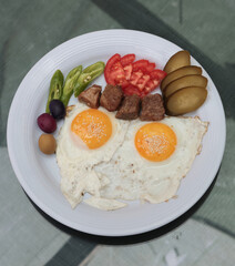 Minimal top view of breakfast with sunny side up eggs, grilled meat cubes, pickles, olives, and tomatoes served on a white plate over a clean glass table, peaceful natural setting ideal for casual