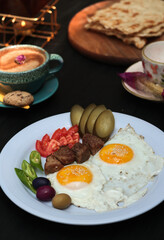 Close-up of a hearty countryside breakfast plate with fried eggs, grilled meatballs, sliced pickles, tomatoes, olives, and cappuccino coffee with artistic foam, beautifully served on a rustic dark woo