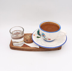 Authentic hot Turkish coffee served in a traditional porcelain cup with intricate floral patterns, accompanied by a glass of water and a chocolate chip cookie, all presented on a rustic wooden tray