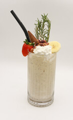 Delicious banana smoothie topped with whipped cream, banana slices, chocolate decorations, strawberries, and rosemary sprigs, served in a tall clear glass with a black straw, captured against a clean