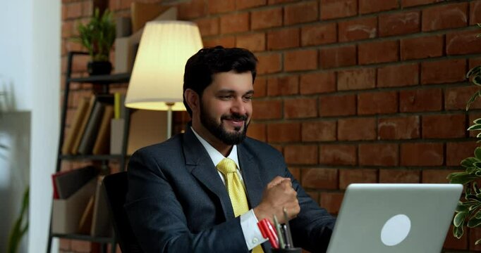Happy Indian Asian handsome young businessman or CEO in formal corporate attire with beard, working on laptop computer in cozy modern office with terracotta brick wall