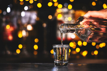 Bartender pours vodka in glass on black bar counter background with lights