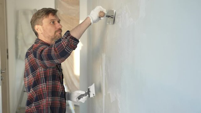 Male construction worker wearing protective gloves, spreading plaster evenly on wall surface using putty knife, renovating interior with professional precision and smooth technique