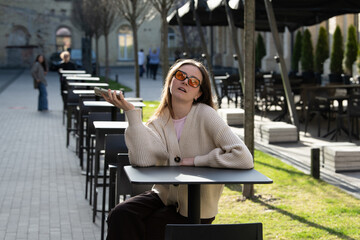 A young woman with sunglasses relaxes at an empty outdoor cafe. She is seated at a table, smiling and holding her phone, surrounded by trees and distant people