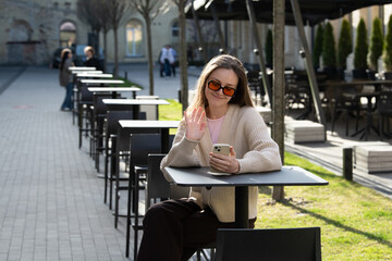 A woman sits at a table in an outdoor cafe on a sunny day. She is wearing sunglasses and waving while looking at her smartphone, surrounded by empty chairs and patrons in the background