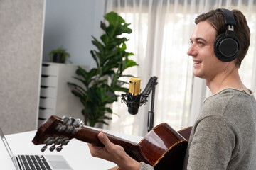 Host channel in smart singer with smiling face, playing guitar along singing, broadcasting on social media channel, wearing headphones to record video streamer at modern studio recording. Pecuniary.