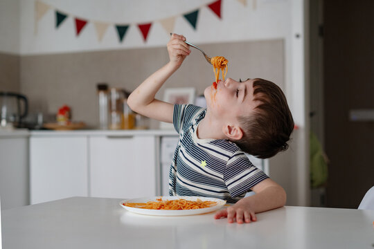 A charming boy eats spaghetti with tomato bolognese. A happy child eats freshly prepared healthy food indoors - Powered by Adobe