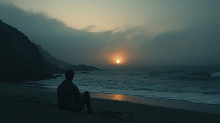 Fototapeta premium A solitary figure gazes at the sunset by the ocean, waves gently rolling in under a misty sky, capturing a moment of peace.
