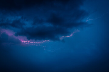 A jagged lightning bolt cuts through dark storm clouds in a vivid blue night sky, creating a...