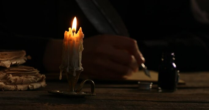 Woman writing with fountain pen at wooden table in darkness, focus on burning candle in candlestick