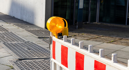 A construction barrier with a bright yellow warning light on a city sidewalk, signaling ongoing...