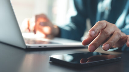 Business woman using mobile phone and working on laptop computer on table, at office, closeup, backlit. Woman busy working on laptop, using smartphone, surfing the internet, social networking