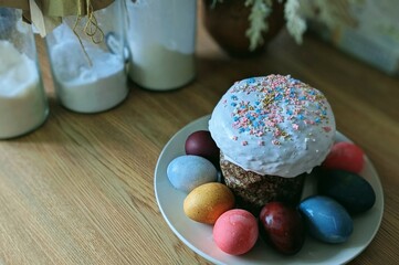 Еaster setting on a wooden table. traditional Easter cake (Kulich) sits on a white plate. The cake with white icing topped with colorful sprinkles. Surrounding the cake are several dyed Easter eggs 