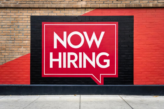 A now hiring banner displayed on a brick wall for job recruitment employment opportunity and vacancy advertisement with bold text and red background for business hiring communication
