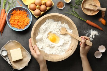 Woman making dough for carrot cake at dark textured table with ingredients, top view