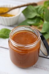 Tasty curry sauce in glass jar, powder and basil on white wooden table, closeup