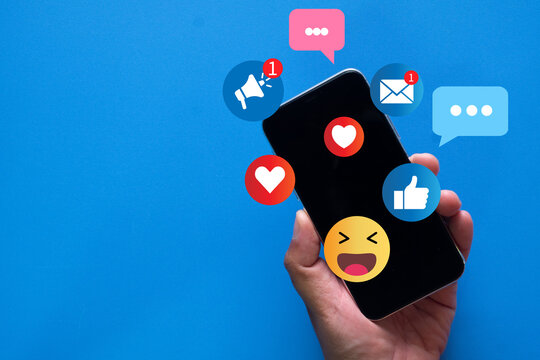 Hand holding smartphone with social media notifications, likes, hearts, and emoji icons on vibrant blue background. Concept of online interaction, social network, and digital communication.