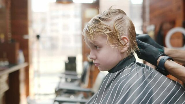 Cute school age boy getting haircut in barbershop. Children hairdresser with professional tools - comb and scissors. Cutting hair for kids. Services of a professional stylist.