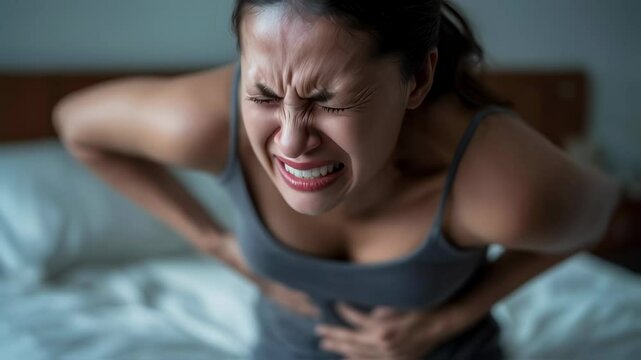 A young woman with dark hair is doubled over in pain, grimacing while holding her abdomen, possibly from cramps or discomfort.