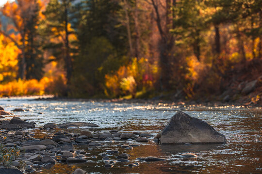 A serene autumn scene along a river in Ouray, Colorado, with vibrant yellow and orange foliage, rocks, and a mountainous backdrop, capturing nature’s seasonal beauty.