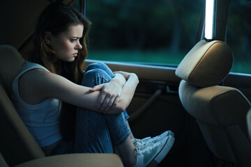 A teenage girl with long hair sitting in a car, reflecting deep emotions while dressed in casual...