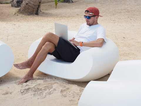 Man in a white t-shirt and sunglasses sits comfortably on white lounge chair at the beach remote working on laptop computer.