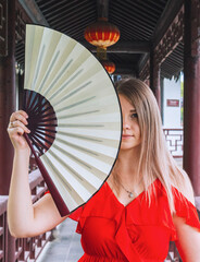 Young woman holds a decorative fan near her face in outdoor area with traditional architecture in the background.