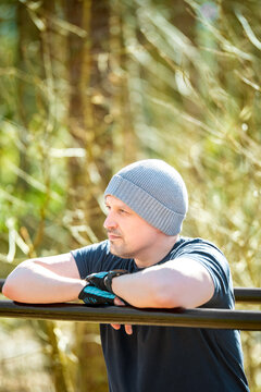 Athletic man resting after outdoor workout, leaning on parallel bars in forest gym. Wearing a beanie and gloves, he looks thoughtful and focused. Fitness, motivation and wellness concept
