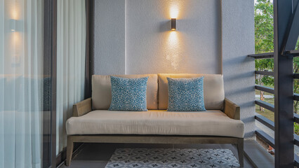 A place to relax on the balcony. A soft beige sofa with patterned cushions stands against the wall. The sconce is lit. Latticed railings. Tulle curtains on the windows. Tiled floor. Philippines.