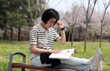 Fototapeta premium A bespectacled woman looking at a book in short-sleeve knitwear on a park bench in warm weather. 